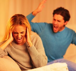 Aggressive Man Shouting At Woman Sitting On Sofa At Home