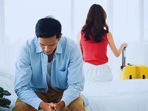 A couple in a bedroom with a tense atmosphere. The man is sitting on the edge of the bed, leaning forward with his hands clasped, appearing stressed or deep in thought. Move Out During a Divorce
